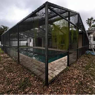 Large dark pool cage over a pool and paver deck, leaf litter on the ground